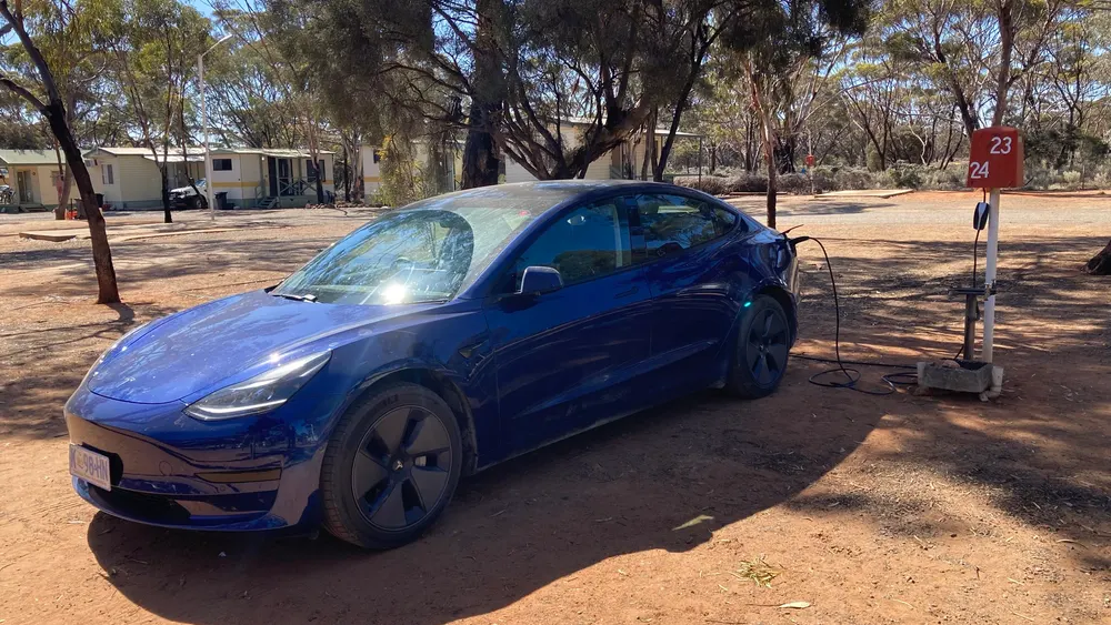 Blaues Tesla Model 3 mit Ladekabel an einer Stromsäule auf einem Campingplatz