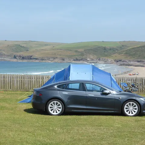 Tesla Model S neben einem Zelt auf einem Campingplatz an der Küste, im Hintergrund Bucht und Sandstrand