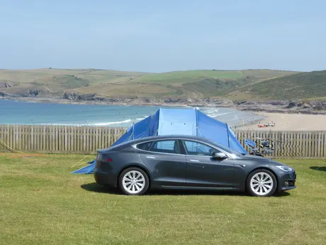Tesla Model S neben einem Zelt auf einem Campingplatz an der Küste, im Hintergrund Bucht und Sandstrand