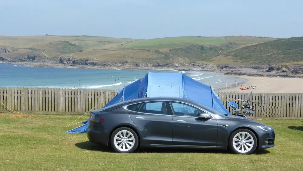 Tesla Model S neben einem Zelt auf einem Campingplatz an der Küste, im Hintergrund Bucht und Sandstrand